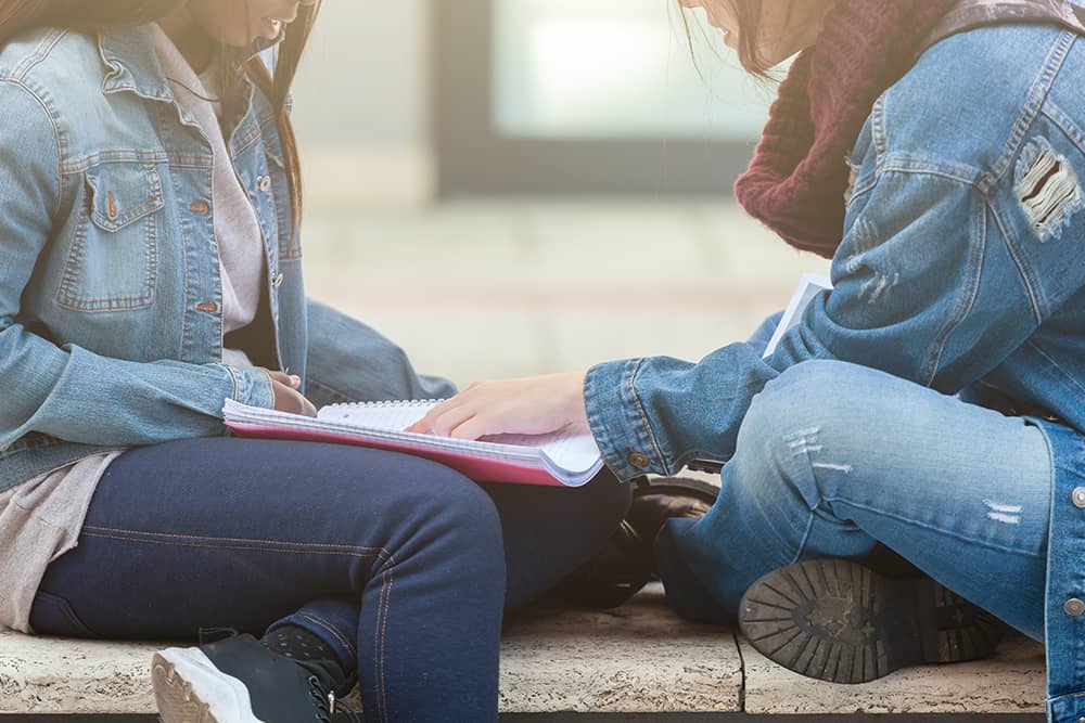 quieres-ser-estudiante-mentor-curso-2024-25 Dos personas sentadas en un banco, una de ellas revisa un cuaderno mientras la otra observa