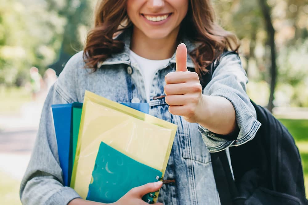 Estudiante sonriente sosteniendo una carpeta y haciendo un gesto de pulgar hacia arriba